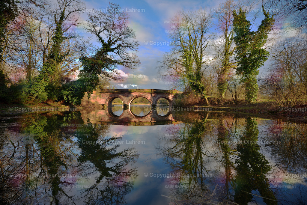 Idyllische Parklandschaft | Eine Parklandschaft im Frühling, in der sich Efeu bewachsene Bäume und eine alte Sandsteinbrücke im Wasser eines See spiegeln, dazu ist ein Blau-Weißer Wolkenhimmel.