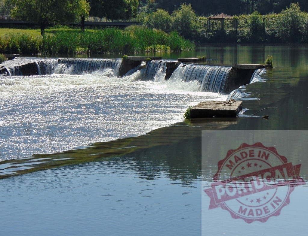 River flowing over concrete dam structures creating weir. Portugal - Norte | Water flowing over a man-made weir on a river, reflecting sunlight and green trees - Realisiert mit Pictrs.com