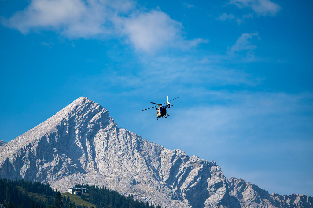 Polizeihubschrauber in den Alpen | Ein Hubschrauber schwebt wie ein kleiner Punkt in der Unendlichkeit eines strahlend blauen Himmels, während im Hintergrund die imposante, schneebedeckte Flanke eines schroffen Berggipfels aufragt. Die Szene vermittelt ein Gefühl von Freiheit und Abenteuer vor der gewaltigen Kulisse der Alpen. - Realisiert mit Pictrs.com