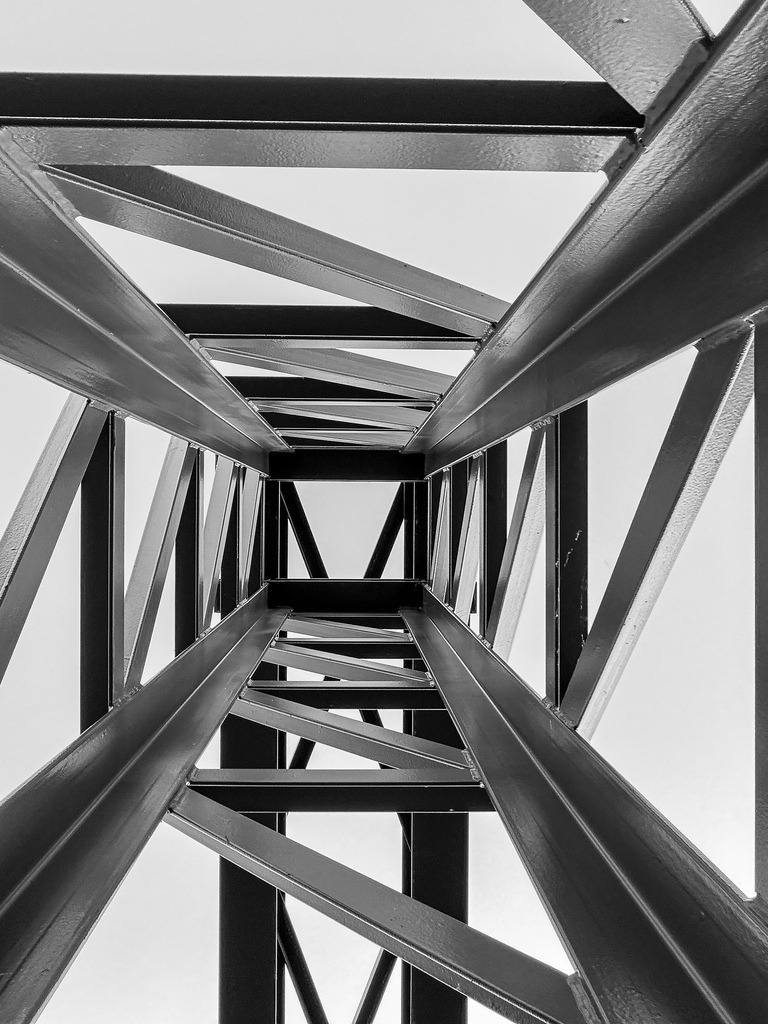 Wandbild Architektur Industrie - Modernes Schwarz-Weiß Poster & Geometrie | Dieses moderne Architektur Wandbild bringt als hochwertiges Poster oder edler Kunstdruck eine kraftvolle, industrielle Atmosphäre in deine Wohnräume. Die faszinierende Schwarz-Weiß-Fotografie zeigt ein geometrisches Stahlskelett in einer beeindruckenden, tiefen Perspektive. Als stilvolle Wanddeko passt diese urbane Fotokunst perfekt zu modernen Einrichtungsstilen und puristischen Lofts im Industrial Design. Wähle jetzt deine Wunschgröße und dein bevorzugtes Material wie Leinwand, Alu-Dibond oder Acrylglas und bestelle diesen architektonischen Blickfang direkt im Shop zu dir nach Hause! - Realisiert mit Pictrs.com