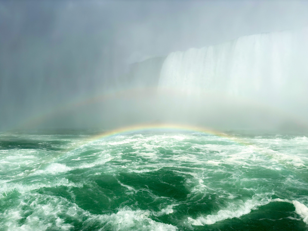 Regenbogenromantik, Kanada | Das Bild zeigt die mächtigen Niagarafälle im Nebel des tosenden Wassers. Ein zarter Regenbogen schwebt über dem smaragdgrünen, aufgewühlten Fluss. Die gewaltige Gischt der herabstürzenden Wassermassen erzeugt im Sonnenschein eine mystische Atmosphäre. - Realisiert mit Pictrs.com