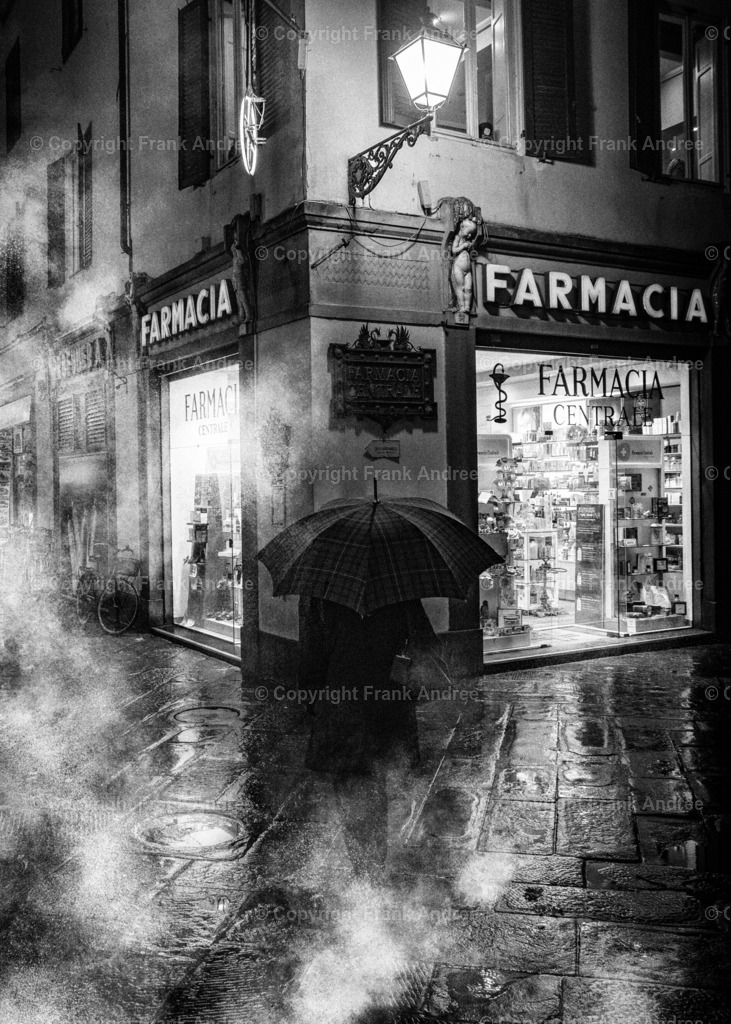 Regen in Lucca  | Eine regnerische Nacht in Lucca Toskana. Eine Frau mit Regenschirm spaziert vor einer beleuchteten Apotheke. Straßenfotografie aus Italien in schwarz weiß.  - Realisiert mit Pictrs.com