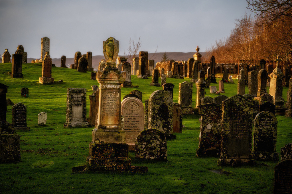 Friedhof Gairloch | Ein weitlaeufiger Friedhof in Gairloch, Schottland, zeigt eine dichte Ansammlung alter, moosbewachsener Grabsteine und monumentaler Denkmaeler. Das warme Abendlicht beleuchtet die Szene von der Seite, betont die Texturen der Steine und wirft lange Schatten ueber das gruene Gras. Die Komposition fuehrt den Blick durch die Reihen der Grabsteine, die von kahlen Baeumen im Hintergrund eingerahmt werden. - Realisiert mit Pictrs.com