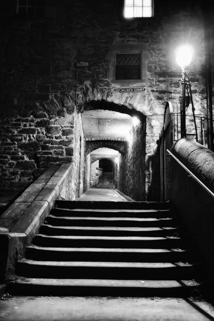 Milne's Court, Edinburgh: Nachttreppe | Eine enge, von Steinmauern gesaumte Gasse in Edinburghs Altstadt fuhrt uber eine Treppe in eine Reihe von bogenformigen Durchgangen. Die Szene ist in Schwarz-Weiss gehalten und wird von einer Strassenlaterne und Fenstern beleuchtet, was eine tiefe Perspektive und ein historisches Ambiente schafft. Diese Komposition betont die architektonische Tiefe und das urbane Erbe der Stadt. - Realisiert mit Pictrs.com