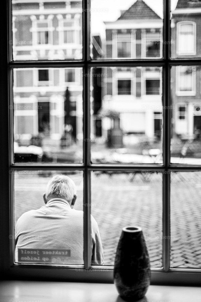 Mann vor dem Fenster | Ein älterer Mann sitzt nachdenklich vor einem Fenster mit Blick auf einen städtischen Platz. Das Bild strahlt stille Ruhe und eine Momentaufnahme vor dem Trubel aus – ideal als Kunstdruck für reflektierende Raumgestaltungen. - Realisiert mit Pictrs.com