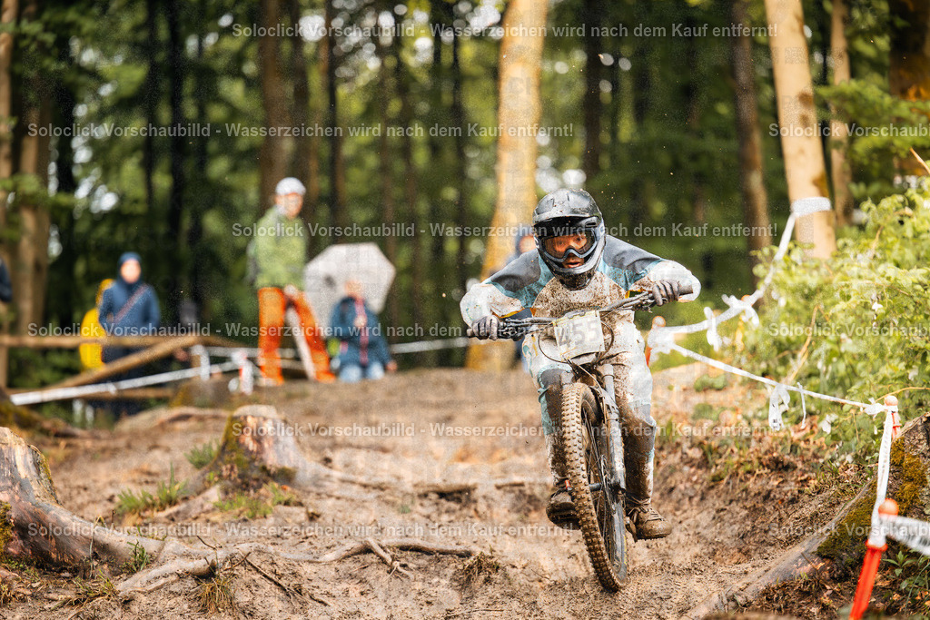 Mountainbike-Action im Wald | Ein Mountainbiker, der mit hoher Geschwindigkeit einen schlammigen, kurvigen Waldpfad durchquert. Er trägt einen weißen Helm mit der Nummer 55 und eine Schutzbrille. Sein Trikot ist mit Schlamm bedeckt. Im Hintergrund sind Zuschauer mit Regenschirmen und Absperrband zu sehen. - Realisiert mit Pictrs.com