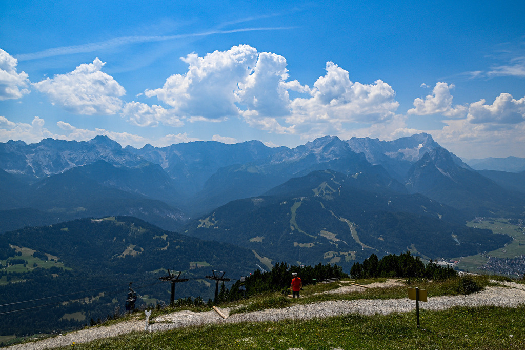 Gipfelmeer über dem Werdenfelser Land | Ein atemberaubender Weitblick vom Wank offenbart die gewaltige Kulisse des Zugspitzmassivs in seiner ganzen Pracht. Der Blick schweift über das weite Tal von der markanten Alpspitze bis hin zur majestätischen Zugspitze, die sich stolz unter einem lebendigen Sommerhimmel mit bauschigen Quellwolken erhebt. Im Vordergrund lädt ein geschwungener Bergpfad zum Innehalten ein, während vereinzelt Wanderer die unendliche Weite und Freiheit der bayerischen Alpen auf sich wirken lassen. Ein Bild, das die Sehnsucht nach alpiner Höhe weckt. - Realisiert mit Pictrs.com