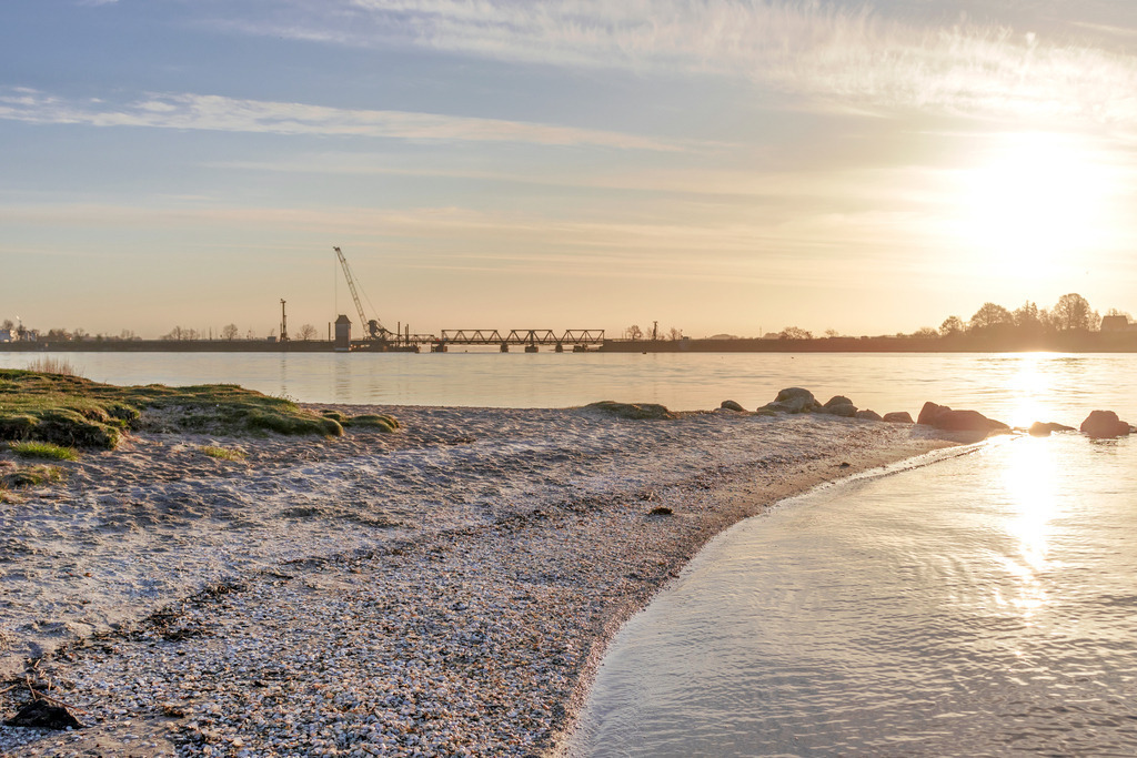 Wandbild: Sonnnenaufgang am Schleistrand Schneiderhaken | Dieses Wandbild im Querformat zeigt einen schönen Sonnenaufgang am Schleistrand Schneiderhaken. Durch die tiefstehende Sonne ergibt sich eine schöne Reflexion auf dem Wasser der Schlei. Im Vordergrund befindet sich der Schleistrand. In der Ferne kann man die Lindaunisbrücke im Gegenlicht erkennen. - Realisiert mit Pictrs.com