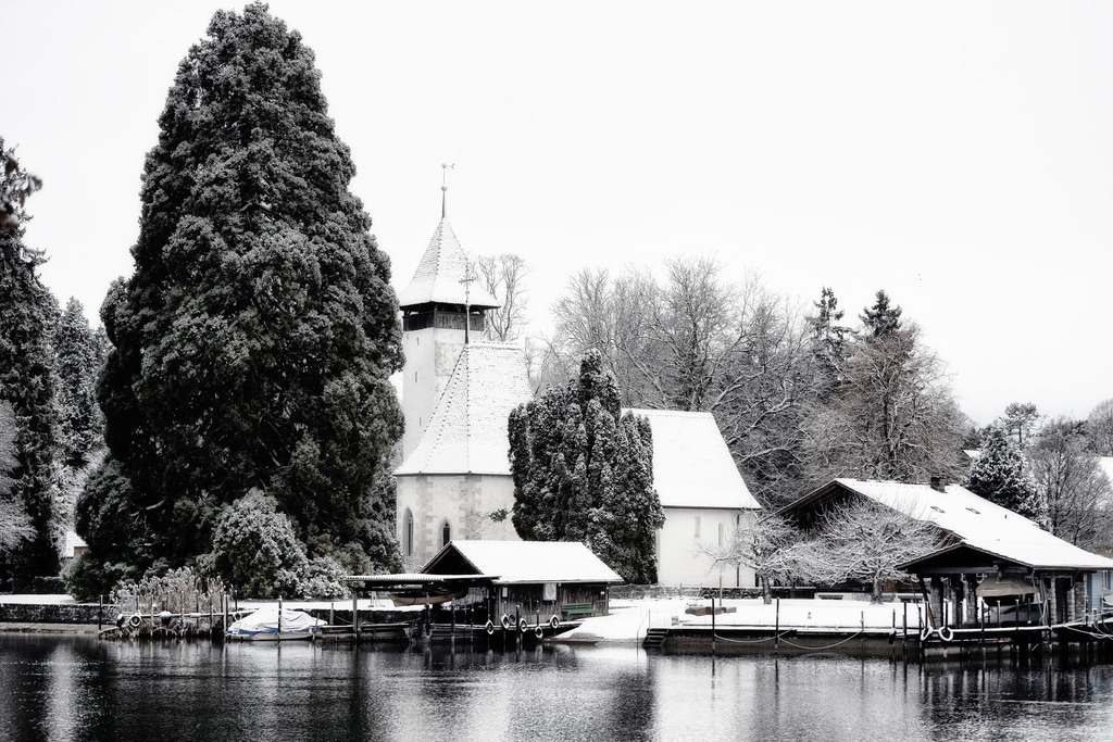 Kirche Scherzligen | Eine schneebedeckte Kirche mit spitzem Turm steht prominent am Ufer des Thunersees, umgeben von dichten, ebenfalls verschneiten Baumen. Im Vordergrund spiegeln sich Bootshauser und das winterliche Ufer im ruhigen Wasser, was eine malerische und ruhige Szenerie schafft.  - Realisiert mit Pictrs.com