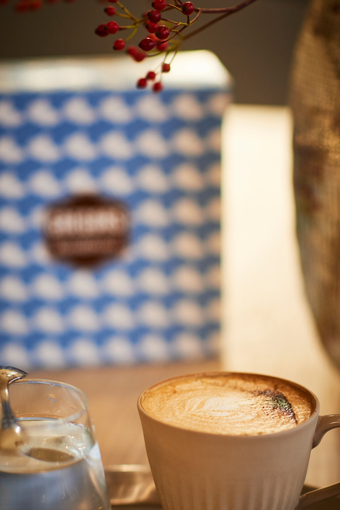 Gregors Konditorei | Wien, Austria - October 20, 2017: Gregors Konditorei, eine Melange mit einem Glas Wasser steht auf dem Tisch, im Hintergrund eine Verpackung und eine Blumenvase. - Realisiert mit Pictrs.com