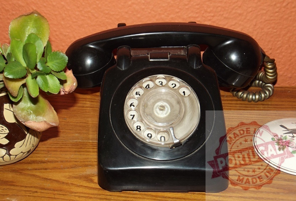 Black rotary phone standing on wooden surface next to plant | Old black rotary phone standing on wooden surface next to plant in a vase and a trinket box - Realized with Pictrs.com