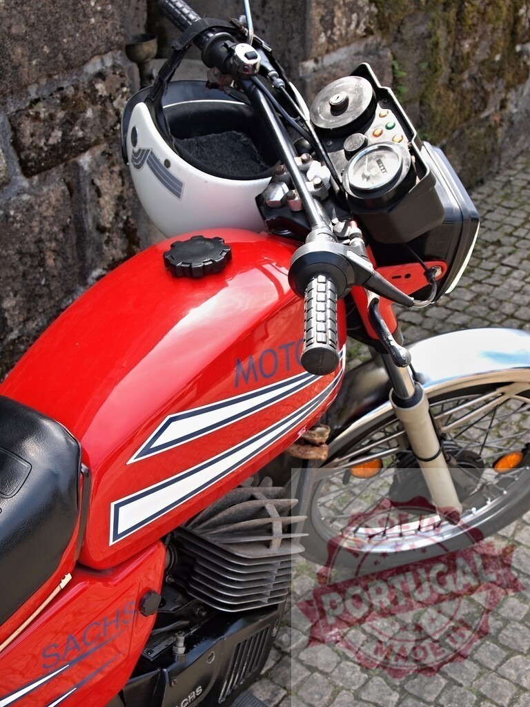 Red Sachs Motorcycle Parked with Helmet on Handlebars | Shiny red Sachs motorcycle parked against stone wall with white helmet hanging from handlebars - Realized with Pictrs.com