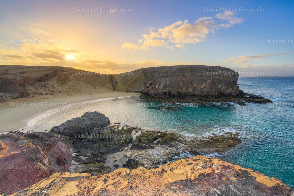 Morgens am Playa de Papagayo auf Lanzarote | Die Sonne geht über den Klippen auf und legt ein erstes, warmes Licht auf die geschwungene Bucht. Die Felsen färben sich langsam von Dunkelrot zu Gold, während das Meer fast lautlos gegen den hellen Sand läuft. In diesem Moment wirkt der Playa de Papagayo wie ein geschützter Aussichtspunkt, an dem der Tag besonders klar und ruhig beginnt. - Realisiert mit Pictrs.com