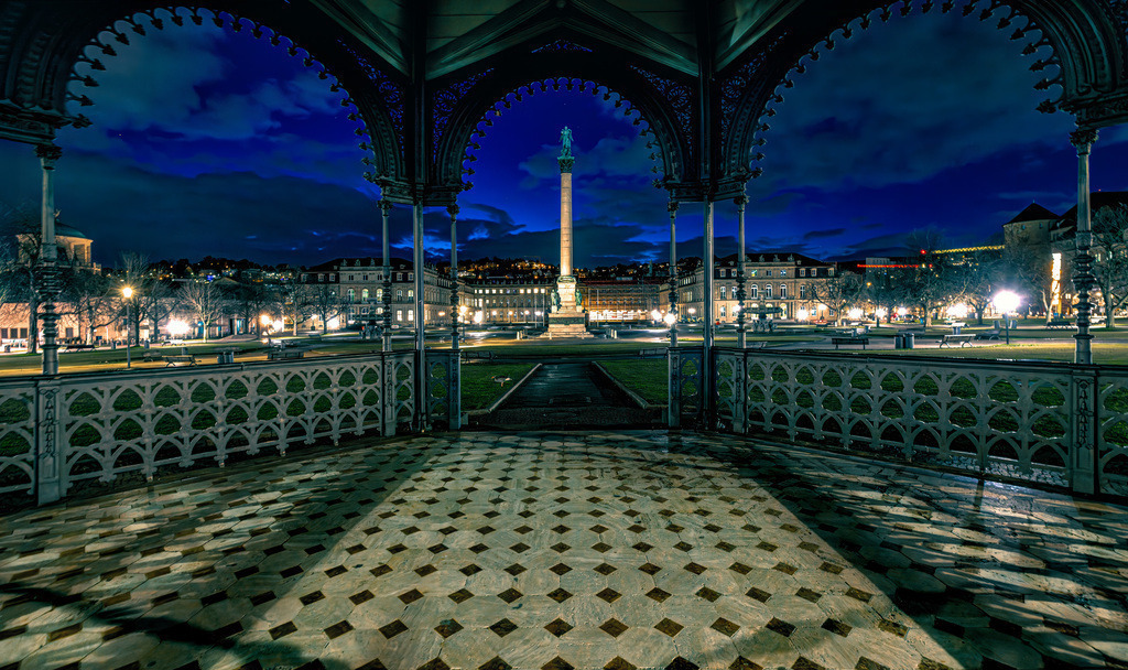 Der Schlossplatz in Stuttgart bei Nacht aus dem Pavillon | Das Bild zeigt den beleuchteten Schlossplatz in Stuttgart bei Nacht, aufgenommen aus einem der historischen Pavillons. Im Zentrum des Platzes ist die rund 30 Meter hohe Jubiläumssäule zu sehen, die 1841 zu Ehren von König Wilhelm I. errichtet wurde. Im Hintergrund befindet sich das Neue Schloss, ein spätbarocker Bau, der heute als Sitz von Ministerien des Landes Baden-Württemberg dient. Die Szenerie ist ein beliebter Treffpunkt und gilt als eines der schönsten Fotomotive der Stadt.  - Realisiert mit Pictrs.com