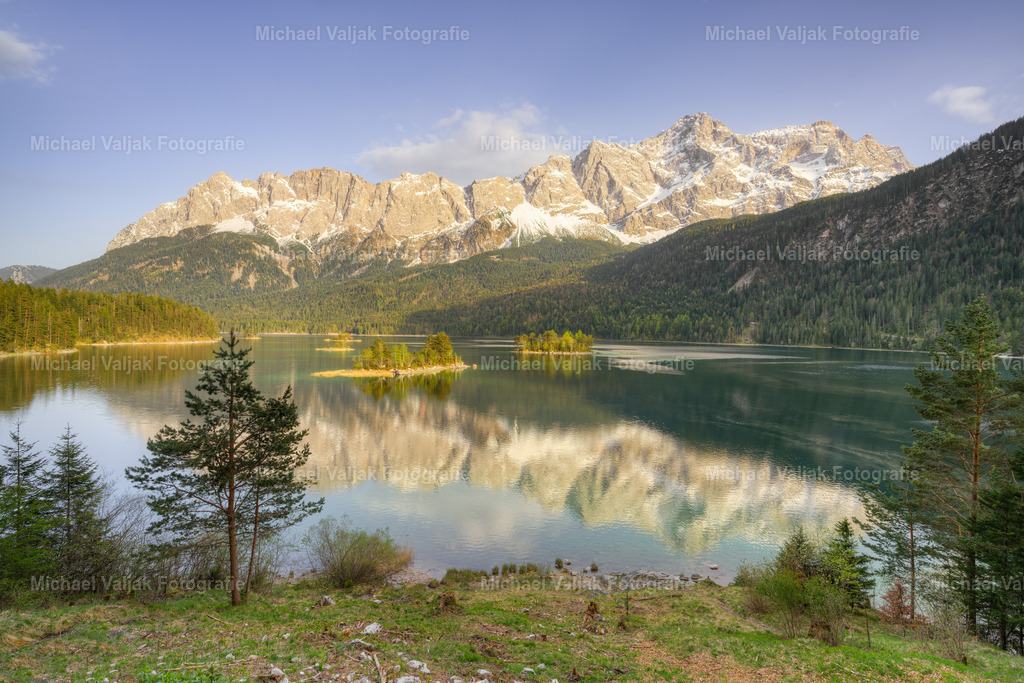 Blick über den Eibsee zum Wettersteingebirge | Aussicht auf das Wettersteingebirge mit der Zugspitze an einem frühen Abend im Frühjahr. Die Berge spiegeln sich im ruhigen See und die Sonne schafft es gerade noch, die Inseln anzuleuchten, bevor sie hinter dem Berg im Rücken verschwindet. - Realisiert mit Pictrs.com
