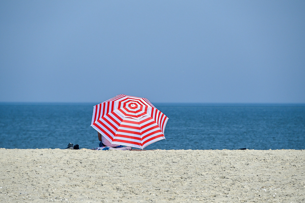 Rückzugsort am Meer | Ein rot-weiß gestreifter Sonnenschirm leuchtet intensiv vor der schier endlosen Kulisse aus tiefblauem Meer und feinem Sandstrand. Unter dem geschützten Dach genießt jemand den perfekten Sommermoment in absoluter Ruhe. Das Zusammenspiel aus klaren Farben, Minimalismus und der Weite des Ozeans weckt sofort die Sehnsucht nach Sonne, Meeresrauschen und purer Entschleunigung am Wasser. - Realisiert mit Pictrs.com