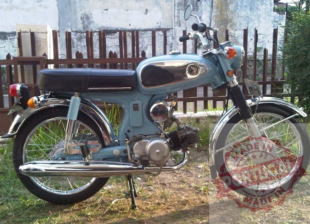 Classic light blue Honda S90 motorcycle parked near a fence | Vintage Honda S90 motorcycle parked on grass near a wooden fence - Realized with Pictrs.com
