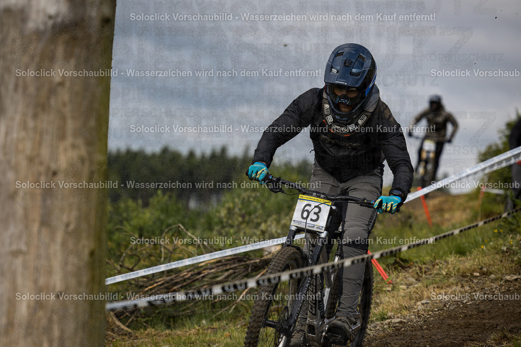 Action im Bikepark Winterberg | Ein Mountainbiker in voller Schutzausrüstung, inklusive Helm und Handschuhen, rast mit seinem Downhill-Mountainbike einen steilen, verschneiten Abhang hinunter. Er trägt eine Startnummer 63 und ist in voller Fahrt zu sehen. Im Hintergrund ist ein weiterer Mountainbiker erkennbar. Die Strecke ist mit roten und weißen Markierungspfosten gesichert. - Realisiert mit Pictrs.com