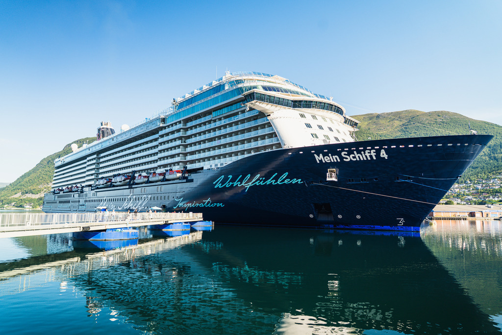 Festgemacht in Norwegen | Nahaufnahme der Mein Schiff 4 an einem schwimmenden Anleger in Nordfjordeid. Das Bild zeigt den markanten Bug des Kreuzfahrtschiffs vor der Kulisse der bewaldeten Bergmassive. Die ruhige Wasseroberfläche spiegelt den dunklen Rumpf und die moderne Architektur des Schiffes wider. - Realisiert mit Pictrs.com