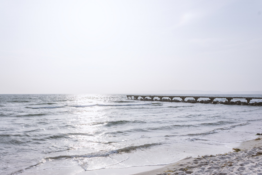 Seebrücke im Morgenlicht – Eckernförde | Die Seebrücke von Eckernförde ragt in das sanft schimmernde Wasser, während feiner Sand und weiche Wellen den Strand in ein stilles Licht tauchen. Die klare Linie der Brücke, das helle Glitzern der Ostsee und die weite, ruhige Küstenlandschaft schaffen ein harmonisches Motiv voller Gelassenheit. Ein Bild, das die friedliche Schönheit der Eckernförder Bucht eindrucksvoll einfängt.