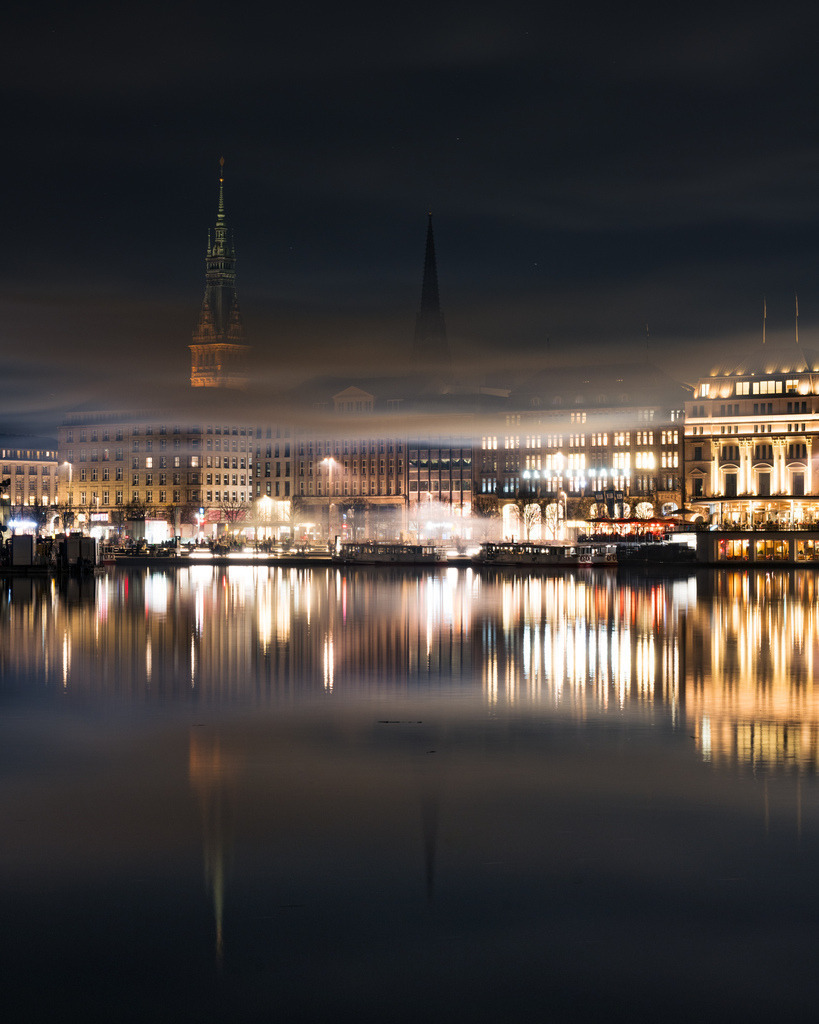 Hamburger Innenstadt im nächtlichen Nebel | Die Lichter der Hamburger Innenstadt spiegeln sich auf der Alster, während tiefe Wolken und leichter Dunst eine geheimnisvolle Stimmung über die Stadt legen. - Realisiert mit Pictrs.com