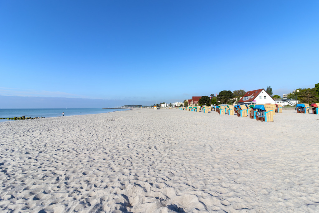 Wandbild: Strandweite am Morgen in Grömitz | Dieses Wandbild zeigt eine weitläufige Strandszene in den frühen Morgenstunden. Der helle Sand erstreckt sich über die gesamte Bildfläche und vermittelt ein Gefühl von Offenheit und Ruhe. Auf der rechten Seite sind zahlreiche bunte Strandkörbe in geordneten Reihen aufgestellt. Im Hintergrund sind mehrere Gebäude zu erkennen, darunter weiße Häuser mit roten Dächern, die dem Bild eine charmante, strandorttypische Note verleihen. Links zieht sich das ruhige Meer bis zum Horizont, wo in der Ferne ein einzelner Spaziergänger am Wasser entlangläuft – ein stiller Moment, bevor der Tag beginnt. Der wolkenlose Himmel in klarem Blau verstärkt die friedliche Atmosphäre. Dieses Motiv eignet sich ideal als Wandbild für maritime Wohnkonzepte – ob als Leinwandbild, Acrylglasbild, Alu-Dibond FineArt Print oder als Akustikbild. Ein stilvoller Akzent für Wohnzimmer, Büro oder Ferienwohnung. - Realisiert mit Pictrs.com