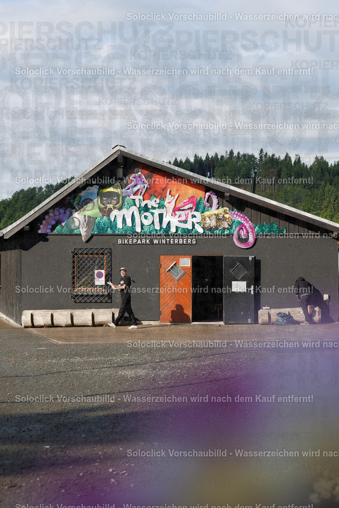 iXS Downhill Cup 2025 - The Mother, Bikepark Winterberg | Das Foto zeigt das Gebäude 'The Mother' im Bikepark Winterberg. Ein Mann in schwarzem T-Shirt und Jeans steht vor dem Eingang und blickt in die Kamera. Das Gebäude ist mit bunten Graffiti verziert und wirkt robust. Im Hintergrund erstreckt sich ein dichter Wald, der die Szene umgibt. Der Himmel ist bewölkt, was auf leicht feuchtes Wetter hindeutet. - Realisiert mit Pictrs.com