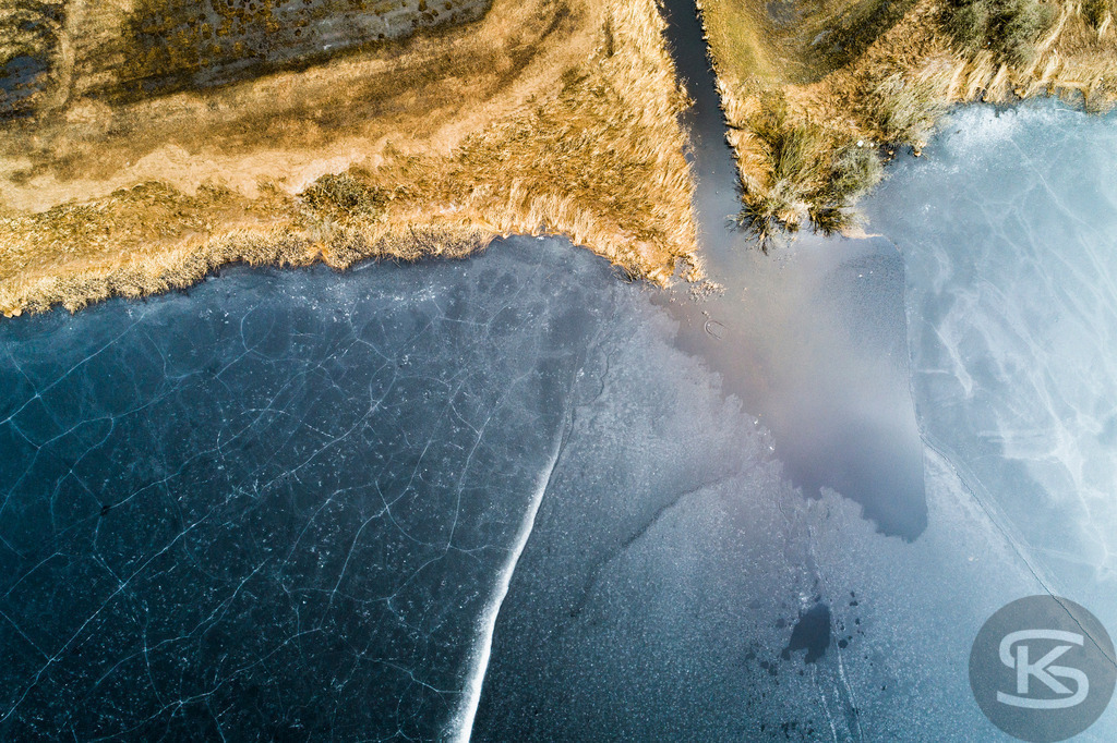 Luftaufnahme: Gefrorener See & Goldenes Ufer im Winter | Hochauflösende Luftaufnahme eines gefrorenen Sees mit interessanten Eisrissen und leuchtend goldenem Schilf am Ufer. Eine stimmungsvolle Winterlandschaft. - Realisiert mit Pictrs.com