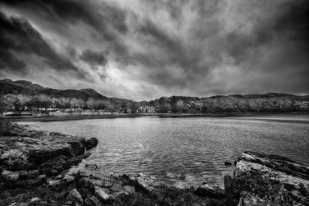 Shieldaig Lodge, Gairloch, Schottland | Eine weite Schwarz-Weiß-Aufnahme zeigt die raue Küstenlandschaft von Gairloch, Schottland, unter einem dramatisch bewölkten Himmel. Im Vordergrund erstrecken sich Felsen und das ruhige Wasser eines Sees oder Meeresarms, während im Mittelgrund ein historisches Gebäude von kahlen Bäumen und Bergen umgeben ist. Die Komposition betont die Tiefe und die kontrastreiche Textur der winterlichen Natur. - Realisiert mit Pictrs.com