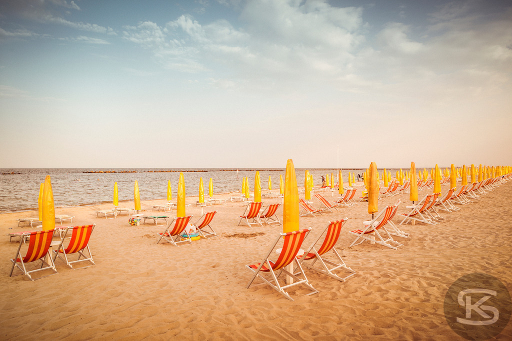 Sonnenverwöhnter Strand mit Liegen an der Adria, Italien | Ein idyllisches Bild eines sonnigen Sandstrandes an der italienischen Adria. Reihen von gelben Sonnenschirmen und rot-weiß gestreiften Liegen säumen den Strand und laden zum Entspannen ein. Die Szene fängt die warme und entspannte Urlaubsatmosphäre perfekt ein, mit ruhigem Meer und weitem Horizont. - Realisiert mit Pictrs.com