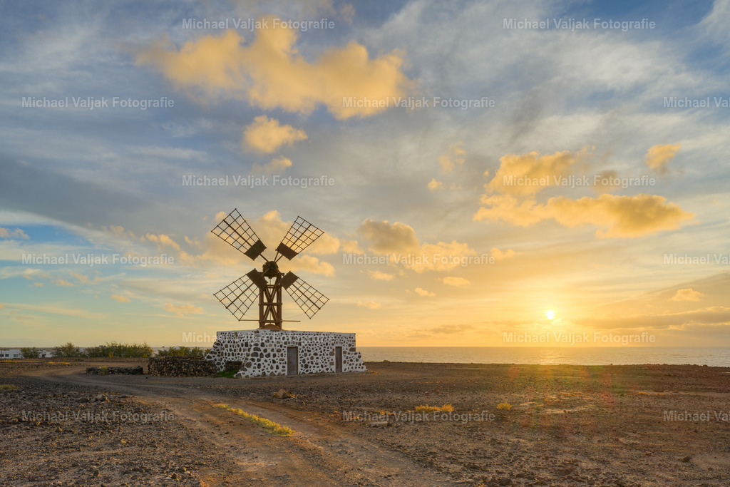 Windmühle in Puerto Lajas auf Fuerteventura | Die Sonne ist bereits aufgegangen und taucht die Windmühle von Puerto Lajas in warmes, direktes Morgenlicht. Die Flügel stehen klar vor dem Himmel, der noch immer von kräftigen Orangetönen durchzogen ist. Die ersten Sonnenstrahlen legen sich über die Landschaft und betonen die Formen der Mühle und des Bodens um sie herum. Ein Moment, in dem der neue Tag mit voller Farbe beginnt und die Mühle wie ein ruhiger Fixpunkt im warmen Licht steht. - Realisiert mit Pictrs.com