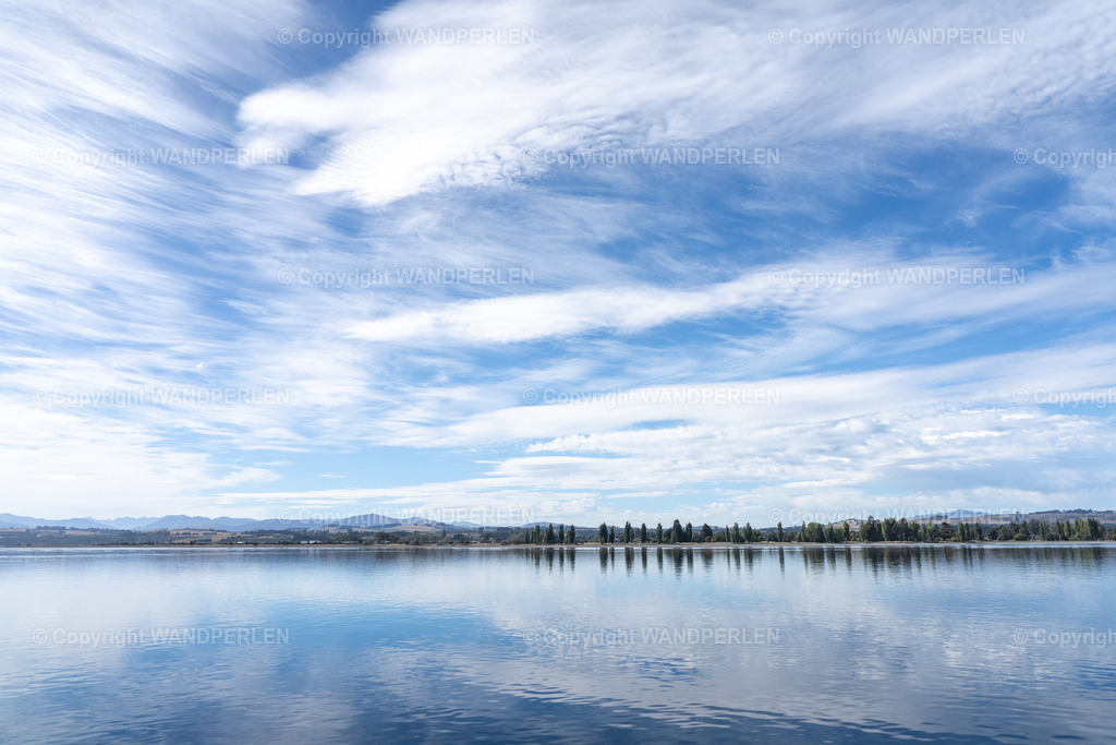 Ein stiller Morgen am Lake Te Anau | Ein ruhiger See, dessen Oberfläche einen weiten, wolkenverhangenen blauen Himmel spiegelt, dominiert die Szene. Eine Reihe von Bäumen und ferne, wellenförmige Hügel bilden den Horizont. Das Bild, aufgenommen bei hellem Tageslicht, zeigt eine friedliche natürliche Landschaft. - Realisiert mit Pictrs.com