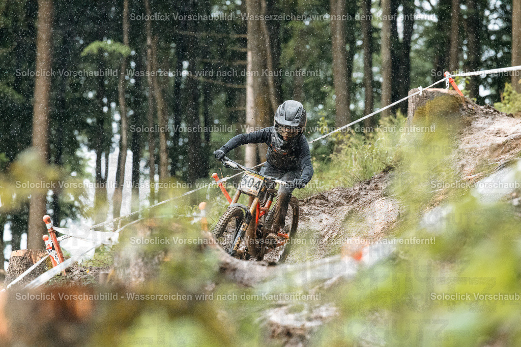 Mountainbike-Abenteuer im Wald | Ein Mountainbiker in voller Ausrüstung rast einen staubigen, kurvigen Trail in einem dichten Wald hinunter. Der Fahrer trägt einen schwarzen Helm und eine schwarze Jacke und Handschuhe. Er trägt die Nummer 504 auf seinem Mountainbike. Der Trail ist mit roten und weißen Markierungen versehen, die den Weg weisen. Der Wald ist dicht bewachsen mit Bäumen und Sträuchern. Es regnet leicht. - Realisiert mit Pictrs.com