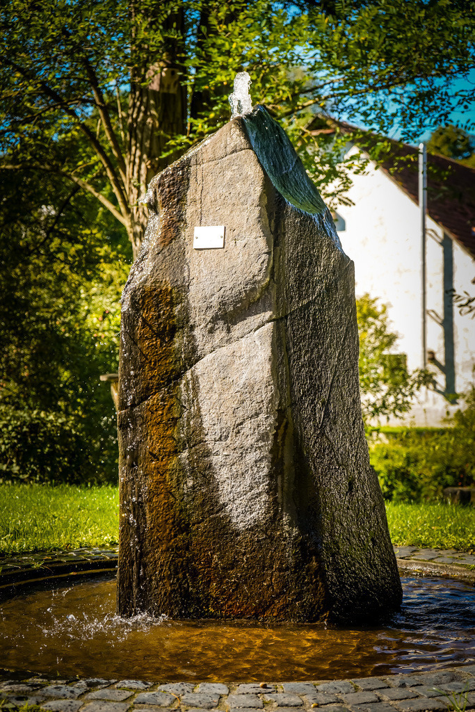 Brunnen - Rinderbügen | An heißen Tagen freut man sich über jede Form der Abkühlung in den Dorfmitten.Auch in Rinderbügen zeigt der Sommer an diesem Wochenende ein letztes Mal was in im steckt. In der sengenden Mittagshitze entfalten die Exponate in der Dorfmitte eine eigene Atmosphäre und die Kunstwerke aus Eisen und Stein werden durch die kontrastspendenden Schatten zum Leben erweckt. Als Besucher spürt man die Liebe und Mühe, die in die Gestaltung und Erhaltung des Platzes fließen. Vielleicht dient dieser Ort als Inspiration für die neuen Dorfmitten in Gedern und Hirzenhain? - Realisiert mit Pictrs.com