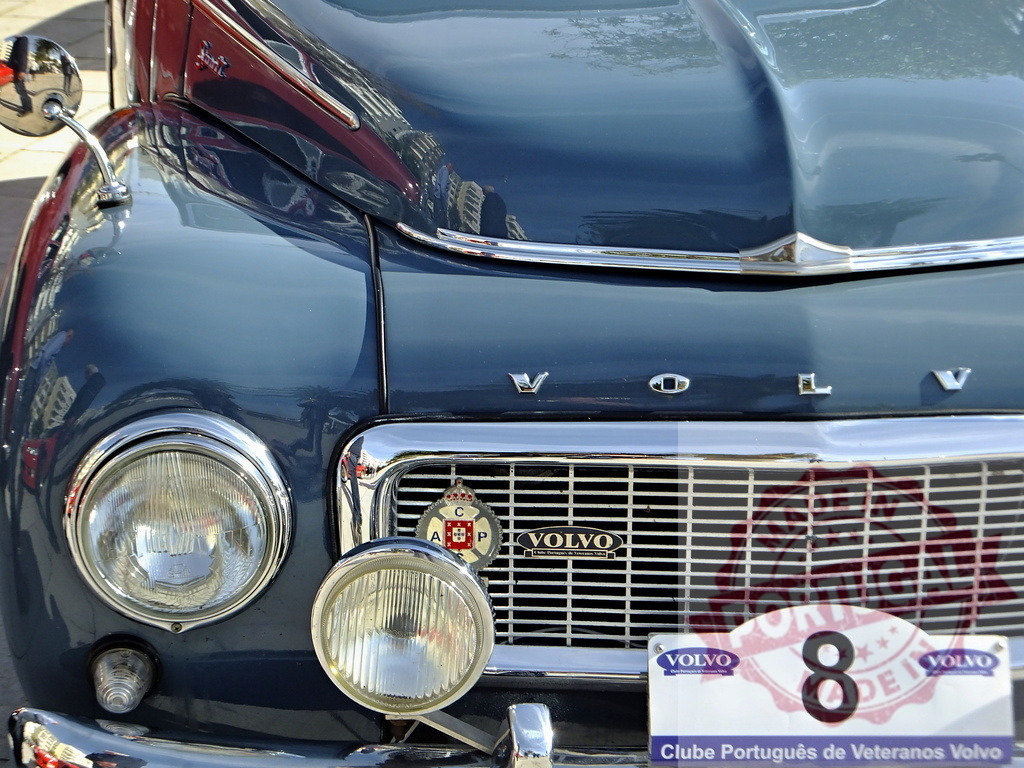 Close-up of Volvo PV444 Front Grill Showing Headlights, Logo, and Clube Português de Veteranos Volvo Badge | Classic Volvo PV444 front grill with original headlights, logo, and Clube Português de Veteranos Volvo badge, reflecting another car - Realisiert mit Pictrs.com
