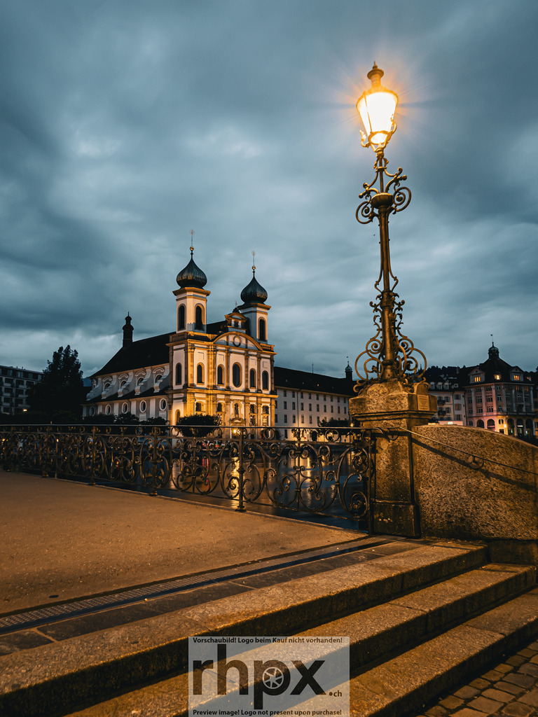 Abend am Rathaussteg | <p><strong>Licht. Symmetrie. Verbindung.</strong></p>
<p>Der beleuchtete Rathaussteg führt den Blick direkt auf das Luzerner Theater unter einem dramatischen Abendhimmel. Die filigranen Verzierungen der Laternen rahmen diesen urbanen Moment perfekt ein und laden zum nächtlichen Flanieren ein.</p>
<p>Wähle unter "Produktauswahl" dein Wunschformat: Vom klassischen Wandbild über Puzzle & Tassen bis zum digitalen Download (z.B. als Handy-Hintergrund).</p>
<br> - Realisiert mit Pictrs.com