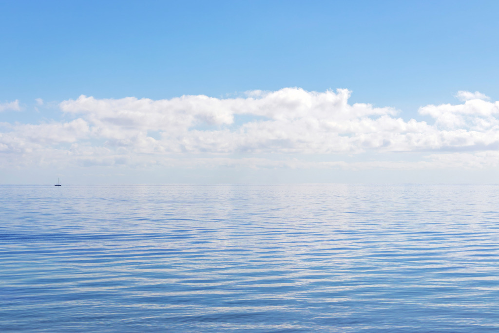 Spiegelung auf ruhiger See – Minimalistisches Meeresbild | Dieses maritime Wandbild zeigt die See in vollkommener Ruhe: Sanfte Wellenstrukturen und leichte Wolken spiegeln sich weich auf der weiten, blauen Wasserfläche. In der Ferne setzt ein kleines Segelboot einen feinen, minimalistischen Akzent, der die meditative Weite des Horizonts perfekt unterstreicht. Die reduzierte Bildsprache macht diese Fotokunst zu einer beruhigenden Ergänzung für moderne Interieurs – ideal als stilvoller Blickfang in Wohnräumen, Büros oder Fewos.