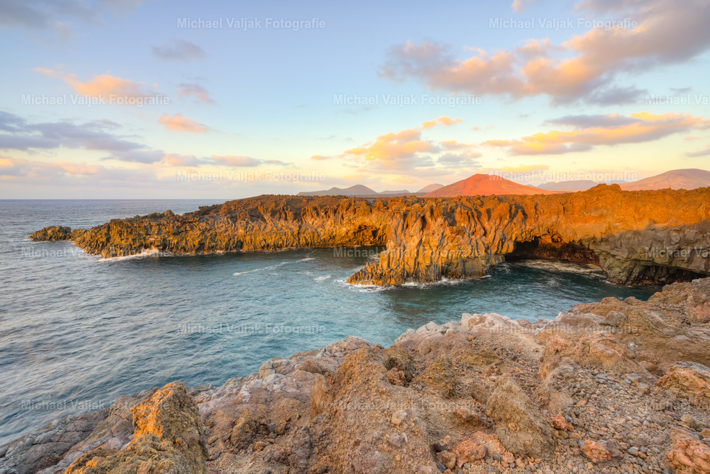 Los Hervideros auf Lanzarote | Die Felsen von Los Hervideros wirken in der warmen Abendsonne fast wie glühende Lava, als würde die Insel für einen Moment ihre vulkanische Herkunft offenbaren. Das Licht streift über die zerklüfteten Klippen, hebt jede Kante hervor und lässt das Meer darunter in tiefem Blau schimmern. Die Brandung des ruhigen Meeres läuft sanft in die Höhlen, während die Sonne langsam sinkt und die Landschaft in ein ruhiges, goldenes Finale taucht. Ein Augenblick, der die raue Kraft Lanzarotes mit einer überraschenden Sanftheit verbindet. - Realisiert mit Pictrs.com