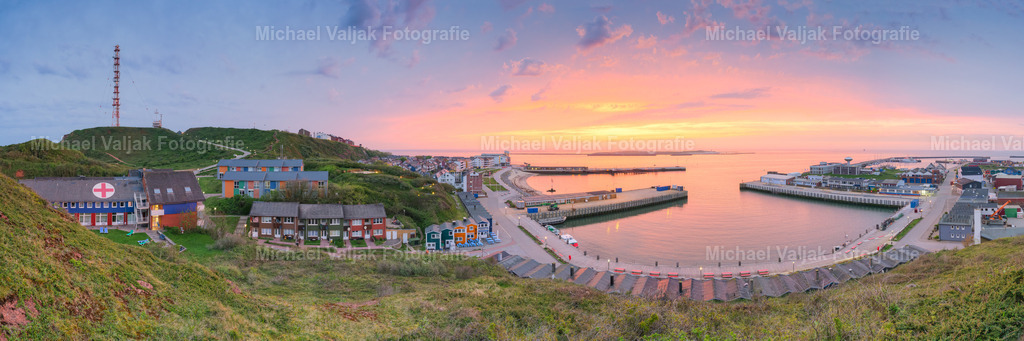 Panoramablick auf Helgoland bei Sonnenaufgang | Das Panorama zeigt Helgoland in der frühen Morgendämmerung. Das Oberland mit seinen Felsen, das Unterland und der Hafen liegen noch im gedämpften Licht, während der Himmel bereits erste warme Farbtöne ankündigt. Die klare Sicht erlaubt einen weiten Überblick über die Inselstruktur, die sich ruhig und kontrastreich vor dem beginnenden Tageslicht abzeichnet. - Realisiert mit Pictrs.com