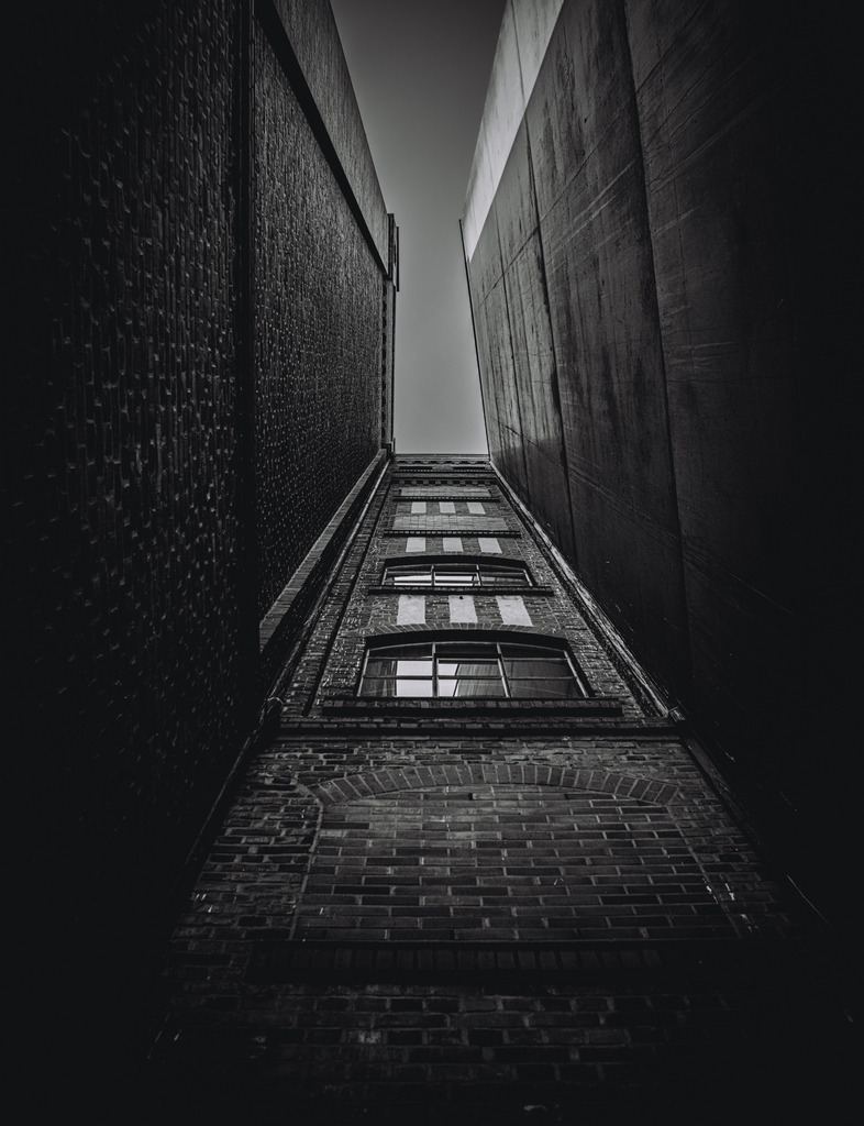 Between Walls and Sky | A dramatic view looking up between towering brick and concrete buildings in Duisburg’s industrial harbor district. The narrow perspective and strong architectural lines draw the eye toward the sky, creating a powerful contrast between urban structure and open space.This fine art architectural photograph captures the raw character of Ruhrgebiet architecture, where historic brick facades meet modern urban geometry. The moody tones and vertical composition emphasize depth, structure, and the quiet atmosphere hidden within the city’s inner courtyards.Perfect as a modern wall art piece for architecture lovers, urban explorers, and collectors of fine art photography. - Realisiert mit Pictrs.com