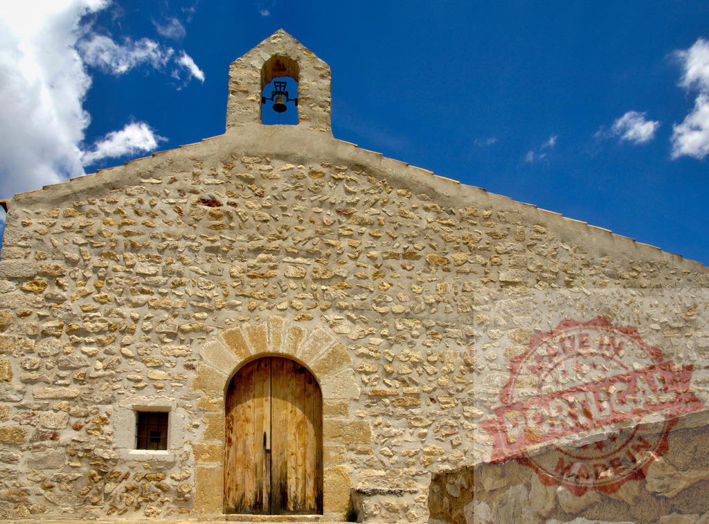 Old stone church facade with bell and blue sky. Spain - Castellon | Old stone church facade with rustic door and bell under a clear blue sky - Realized with Pictrs.com