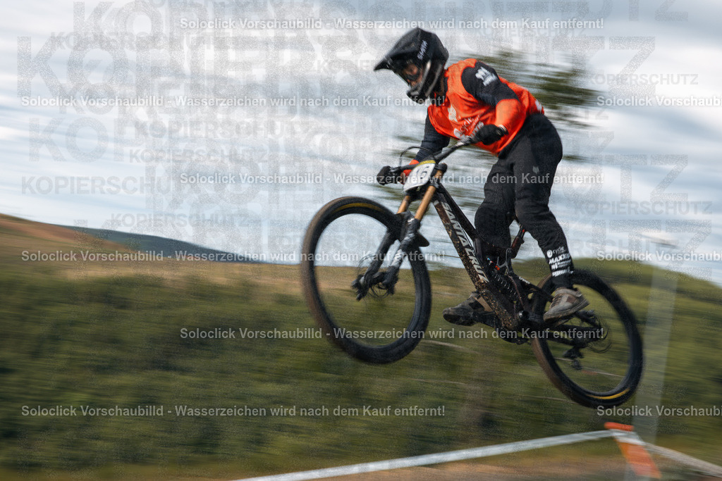 Adrenalin pur: Downhill-Action im Bikepark Winterberg | Ein dynamisches Foto zeigt einen Mountainbiker in voller Aktion während des iXS Downhill Cups 2025 im Bikepark Winterberg, speziell auf der Strecke 'The Mother'. Der Fahrer ist in der Luft, während er eine Sprungrampe überquert. Er trägt einen schwarzen Helm mit orangefarbenen Akzenten und eine orangefarbene Jacke. Sein Mountainbike ist ein Santa Cruz Modell mit auffälligen Komponenten. Die Umgebung besteht aus grünem Gras und einem leicht verschwommenen Hintergrund, der die Geschwindigkeit und die Bewegung des Fahrers betont. - Realisiert mit Pictrs.com