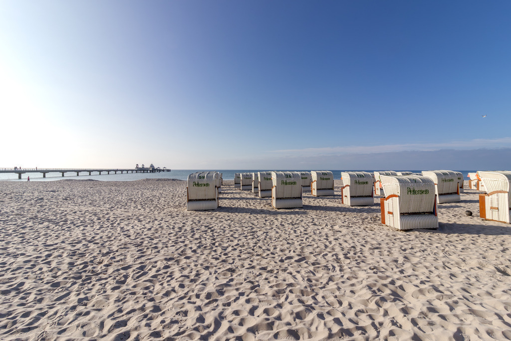Wandbild: Zwischen Sand und Seebrücke – Strandruhe in Grömitz | Dieses Wandbild zeigt eine geordnete Strandszene in Grömitz an einem sonnigen Tag. Auf dem hellen Sand stehen mehrere weiße Strandkörbe in gleichmäßigen Reihen und verleihen dem Bild eine klare Struktur. Im Hintergrund erstreckt sich die Seebrücke von Grömitz weit ins ruhige Meer hinaus. Einige Spaziergänger sind auf der Brücke zu erkennen, und am Ende der Konstruktion steht die markante Tauchgondel, die der Szene Tiefe und architektonischen Charakter verleiht. Der Himmel ist wolkenlos und strahlt in klarem Blau, das Licht fällt weich auf den Sand und die Körbe und schafft eine ruhige, strukturierte Atmosphäre. Die Komposition lebt vom Wechselspiel aus Ordnung und Weite, aus Architektur und Natur – ein stiller Moment der Küstenkultur. Dieses Motiv eignet sich ideal als Wandbild für maritime Wohnkonzepte – ob als Leinwandbild, Acrylglasbild, Alu-Dibond FineArt Print oder als Akustikbild. Ein stilvoller Akzent für Wohnzimmer, Büro oder Ferienwohnung. - Realisiert mit Pictrs.com