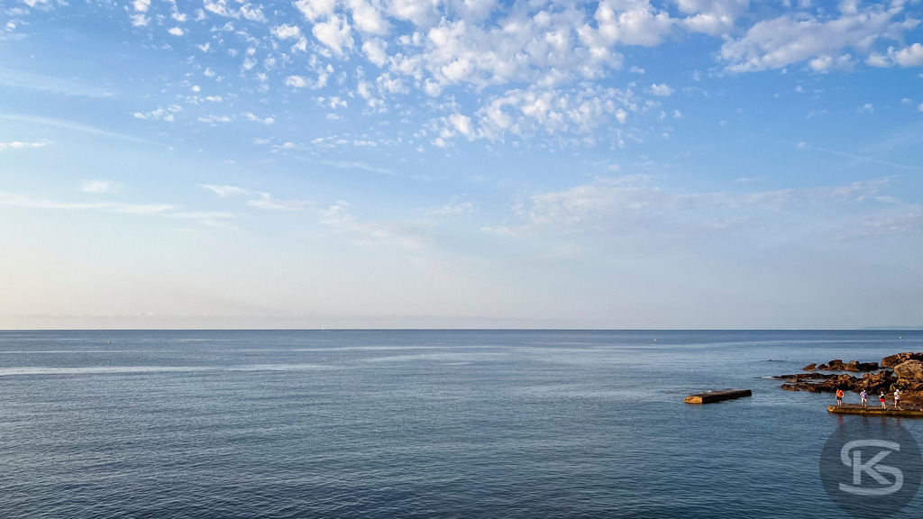 Ruhiges Mittelmeer am Morgen – Küste der Côte d’Azur bei Fréjus, Saint-Raphaël | Weite Panorama-Ansicht des ruhigen, tiefblauen Mittelmeers am frühen Morgen an der Côte d’Azur mit einer felsigen Küste und Personen auf einem Steg unter leicht bewölktem Himmel. - Realisiert mit Pictrs.com