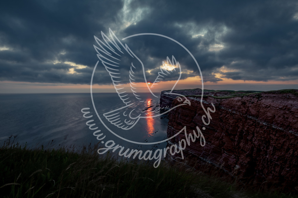 sunset over the red cliffs - germany | Ein dramatischer Abendhimmel über den ikonischen roten Klippen von Helgoland. Die Sonne bricht durch die schweren Wolken und zieht eine leuchtende Reflexion über das ruhige Meer – ein Moment voller Ruhe, Tiefe und atmosphärischer Kraft. Dieses Fine-Art-Landschaftsbild verbindet natürliche Dramatik mit minimalistischer Klarheit und spiegelt die besondere Stimmung der Nordseeinsel im charakteristischen Grumagraphy-Landscape-Look wider.Ein hochwertiger Premium-Print für Wohnräume, Büros und alle Orte, die eine ruhige, moody und zugleich kraftvolle Naturstimmung tragen sollen.