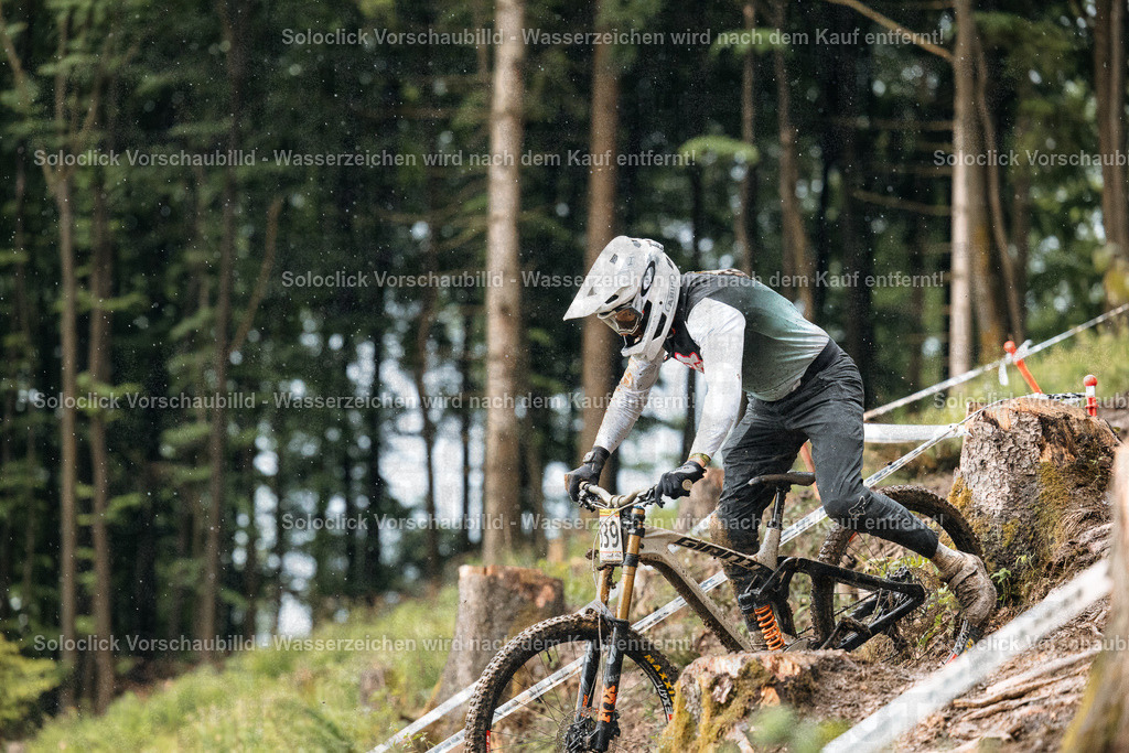 Downhill-Mountainbiker im Regen | Ein Mountainbiker in voller Ausrüstung rast einen steilen, bewaldeten Hang hinab. Er trägt einen Helm, eine Schutzbrille, Handschuhe und spezielle Mountainbike-Kleidung. Sein Fahrrad ist ein Downhill-Mountainbike mit Federgabel und breiten Reifen. Der Hintergrund besteht aus dichtem Grünwald und einem bewaldeten Hang. Es regnet leicht, was die Szene feucht und dynamisch erscheinen lässt. - Realisiert mit Pictrs.com