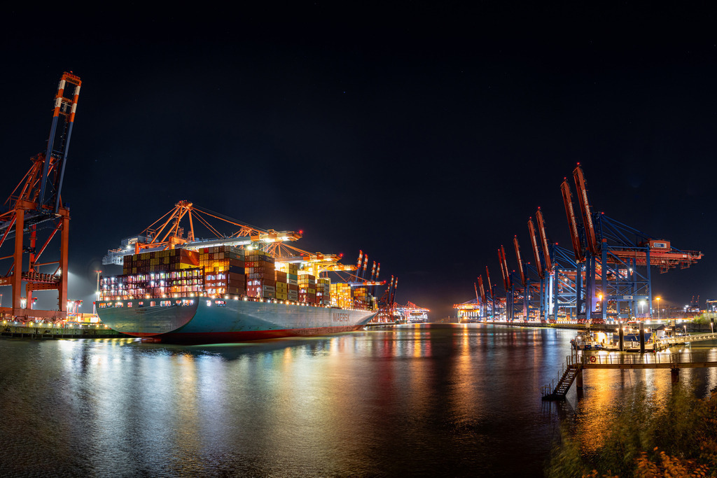 Nachts am Burchardkai Nr.4 | Das Bild zeigt eine nächtliche Szenerie im Hamburger Hafen am Burchardkai, wo mehrere riesige Containerschiffe angedockt sind. Die Schiffe erheben sich majestätisch in die Dunkelheit und werden von einer Flut von Lichtern beleuchtet, die vom Hafen und von den Schiffen selbst ausgehen. Das Bild fängt die Hektik und den Puls des Hafens in der Nacht ein und vermittelt einen Eindruck von der enormen Größe und dem Einfluss dieses wichtigen Handelszentrums.