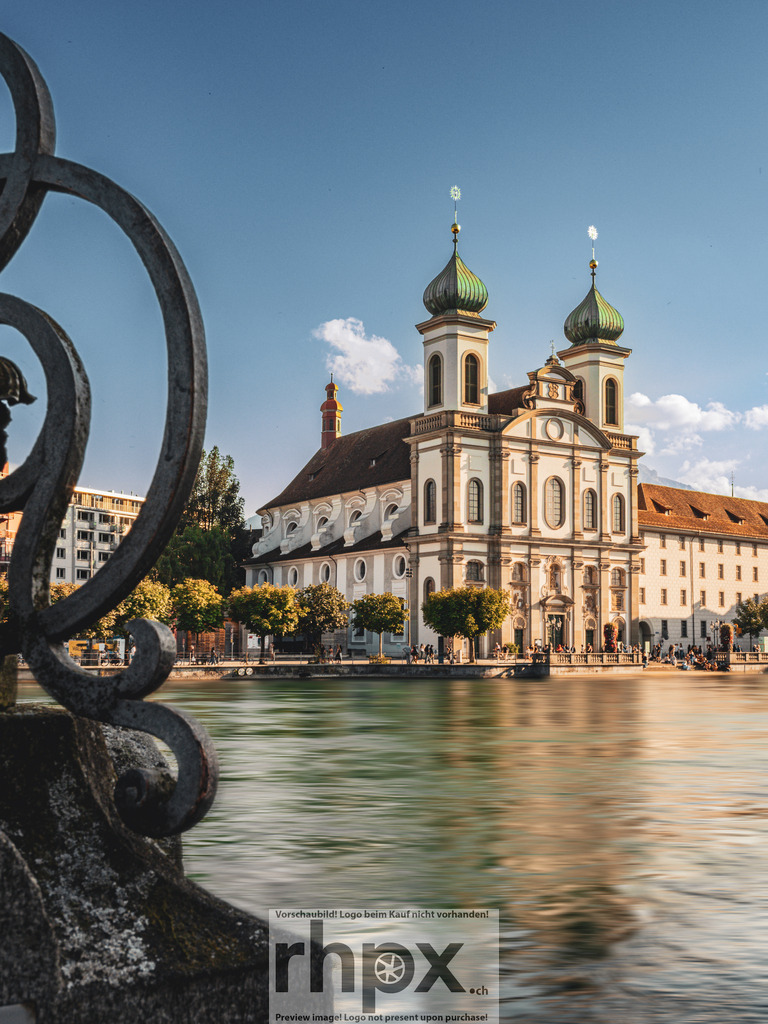 Jesuitenkirche Luzern | <p><strong>Barock. Fluss. Eleganz.</strong></p><p>Die prachtvolle Fassade der Jesuitenkirche leuchtet im warmen Abendlicht an der Reuss. Durch das kunstvolle Eisenwerk im Vordergrund öffnet sich der Blick auf dieses Wahrzeichen von Luzern, während das Wasser die sanfte Spiegelung der Türme trägt. Eine harmonische Verbindung von historischer Architektur und fliessender Bewegung.</p><p>Wähle unter "Produktauswahl" dein Wunschformat: Vom klassischen Wandbild über Puzzle & Tassen bis zum digitalen Download (z.B. als Handy-Hintergrund).</p> - Realisiert mit Pictrs.com
