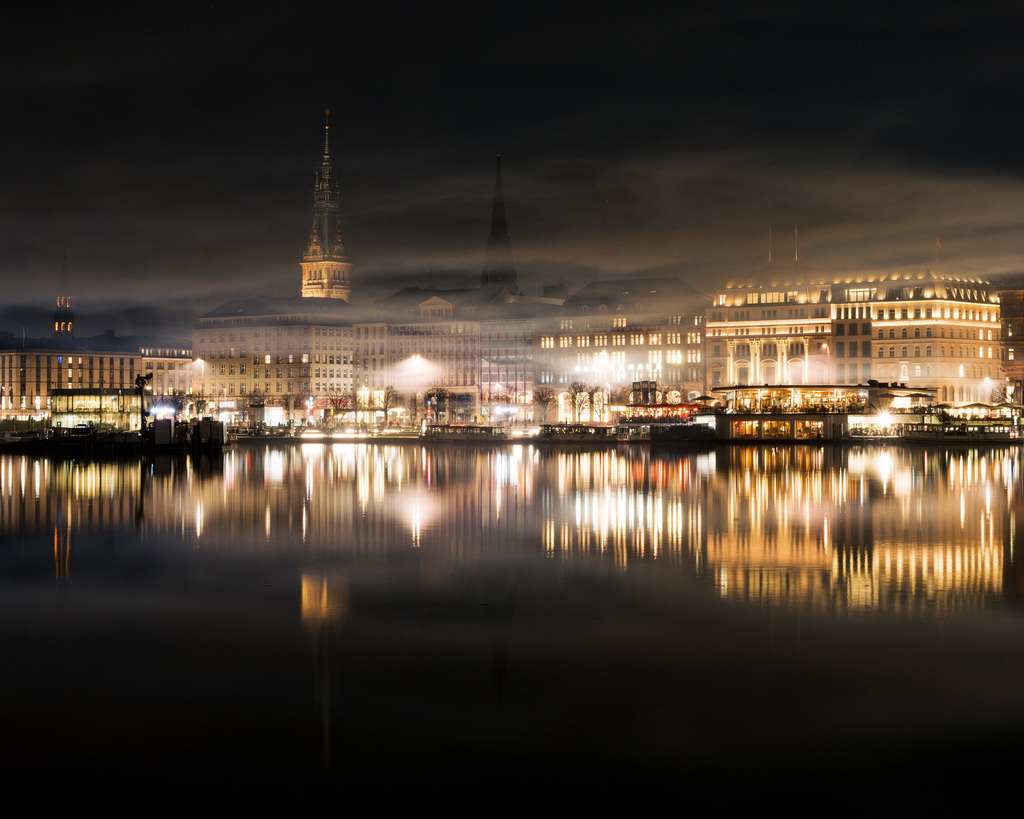 Nächtliche Alsterlichter in Hamburg | Die beleuchtete Hamburger Innenstadt erstreckt sich entlang der Alster. Warme Lichter und ihre Reflexionen treffen auf einen dunklen, atmosphärischen Nachthimmel. - Realisiert mit Pictrs.com