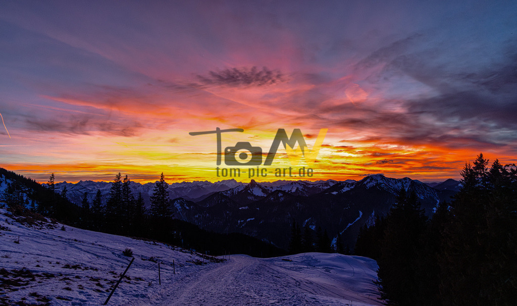 Sonnenaufgang auf dem Wallberg am Tegernsee | Das Bild zeigt einen malerischen Sonnenauf- oder Sonnenuntergang über einer verschneiten Berglandschaft. Der Himmel ist in leuchtenden Farben von Gelb, Orange, Rosa und Violett gehalten.Die Szene zeigt eine Bergkette, die sich silhouettenhaft gegen den farbigen Himmel abzeichnet.Im Vordergrund ist eine schneebedeckte Piste oder ein Weg zu sehen, der von Bäumen gesäumt ist.Ähnliche Sonnenaufgangsansichten sind beliebte Motive am Wallberg oder Walmendingerhorn in den Alpen. - Realisiert mit Pictrs.com